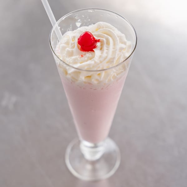Pink milkshake with whipped cream and a cherry on top, served in a tall glass with a straw.