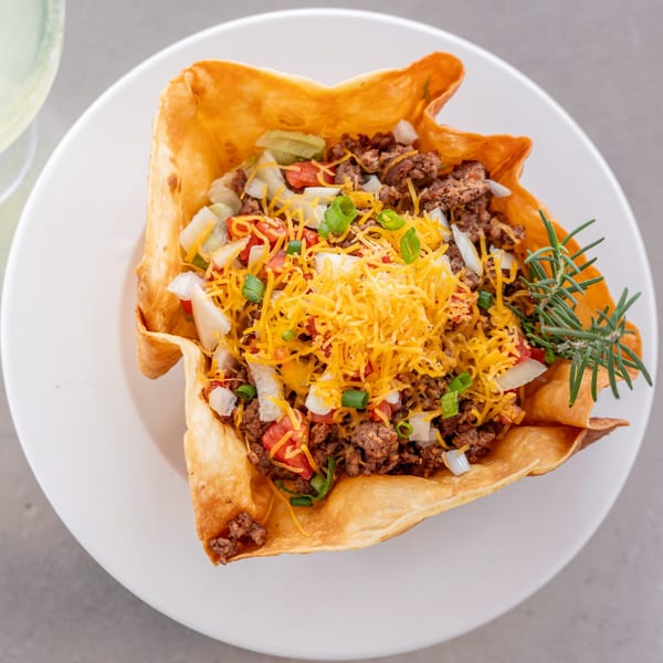 A taco salad in a crispy tortilla bowl, topped with ground beef, cheese, tomatoes, onions, and green onions.