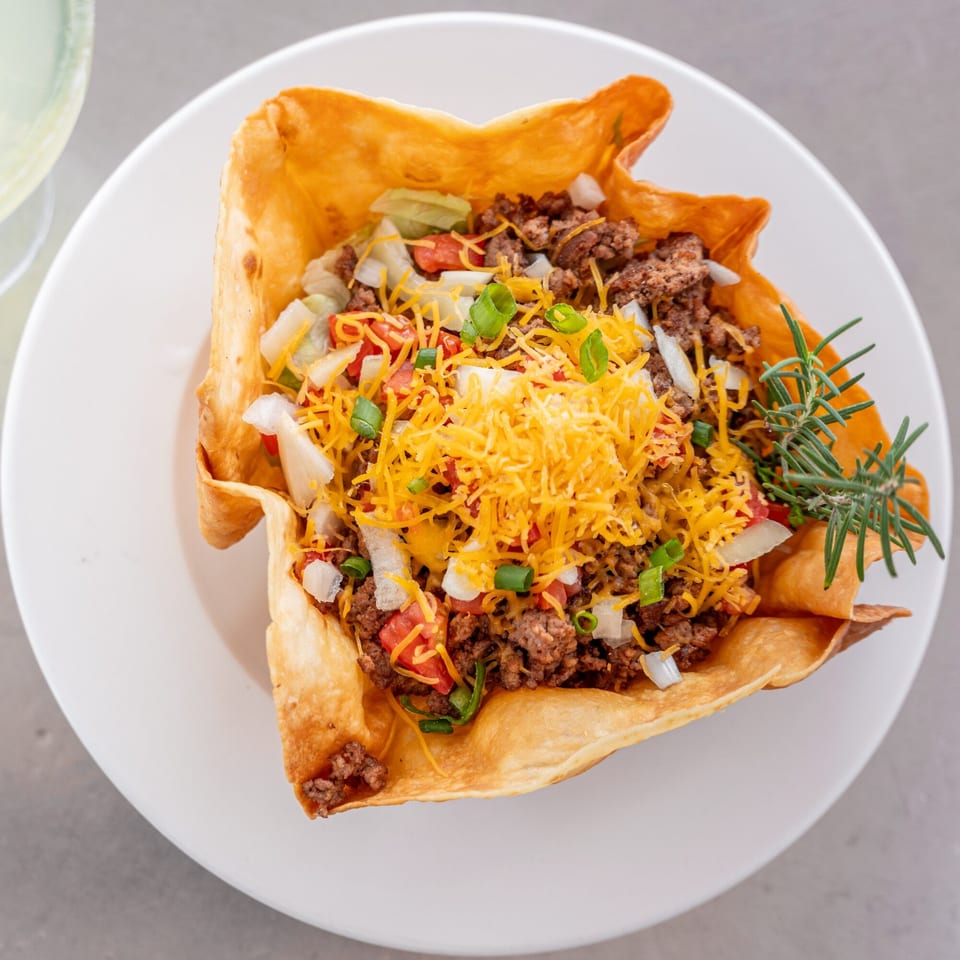 A taco salad in a crispy tortilla bowl, topped with ground beef, cheese, tomatoes, onions, and green onions.