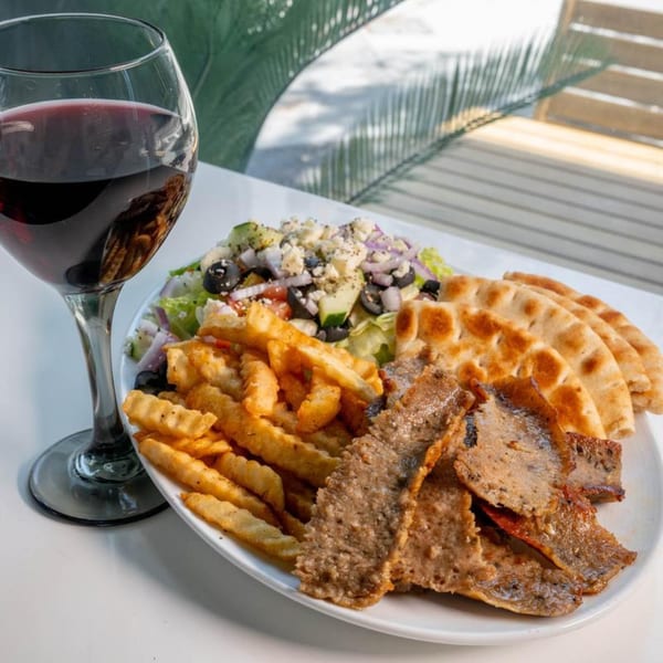 A plate of gyro meat, fries, Greek salad, pita bread, and a glass of red wine.